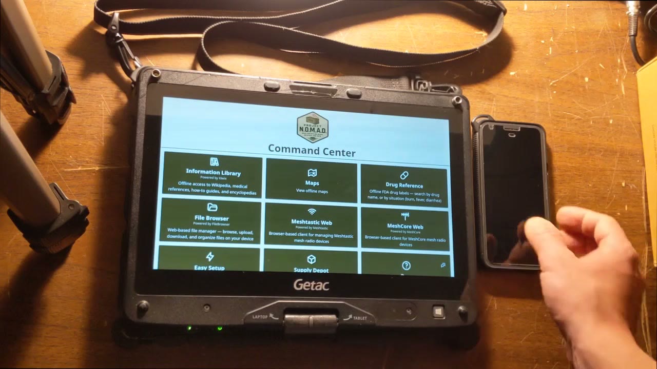 A Getac rugged convertible laptop on a wooden table running the Project NOMAD Command Center, showing tiles for Information Library, Maps, Drug Reference, File Browser, Meshtastic Web and MeshCore Web, with a phone beside it