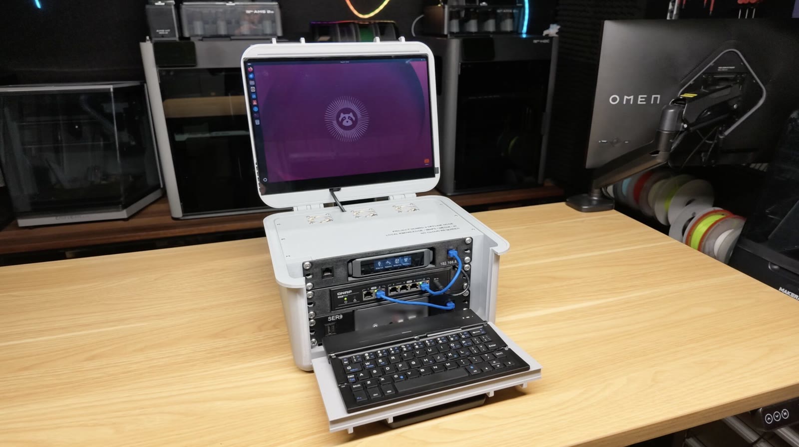 Michael Klements’ 3D printed portable homelab open on a desk: a printed white case holding a 3U 10-inch rack with a network switch and mini PC, a touchscreen in the hinged lid, and a folding keyboard tray at the front